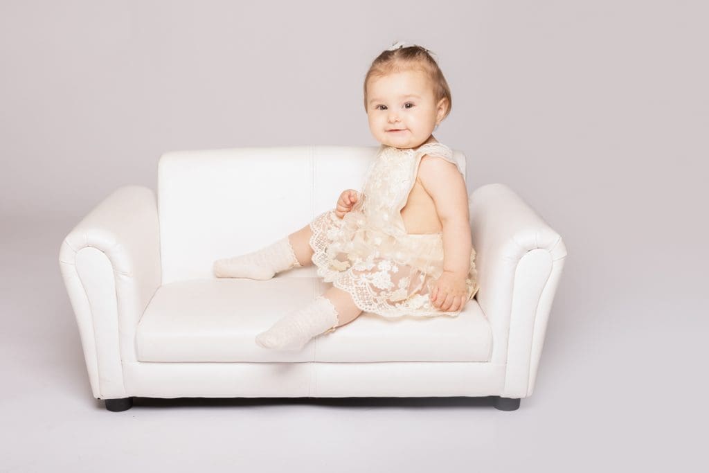 Baby girl sitting sideways on a small white sofa during a professional baby photoshoot in Birmingham.