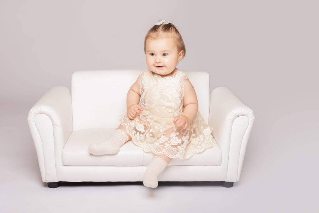 Baby girl sitting on a small white sofa wearing a lace dress during a professional baby photoshoot in Birmingham.