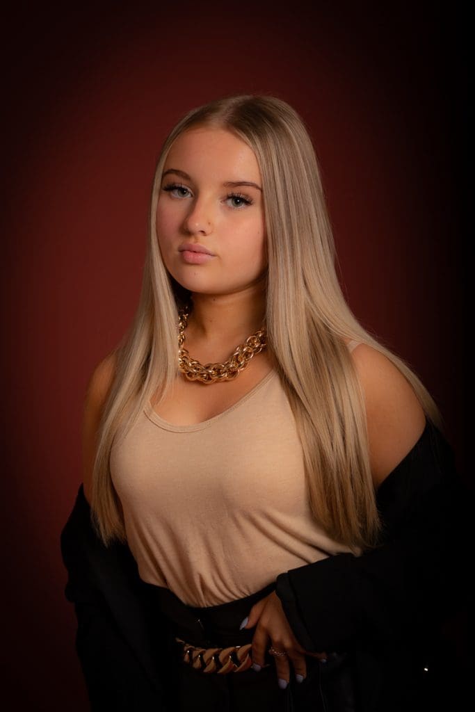 Young woman with long, straight blonde hair stands against a dark red backdrop. Shes wearing a beige top and a chunky gold necklace, with a black jacket draped over her shoulders. Her expression is calm and confident.