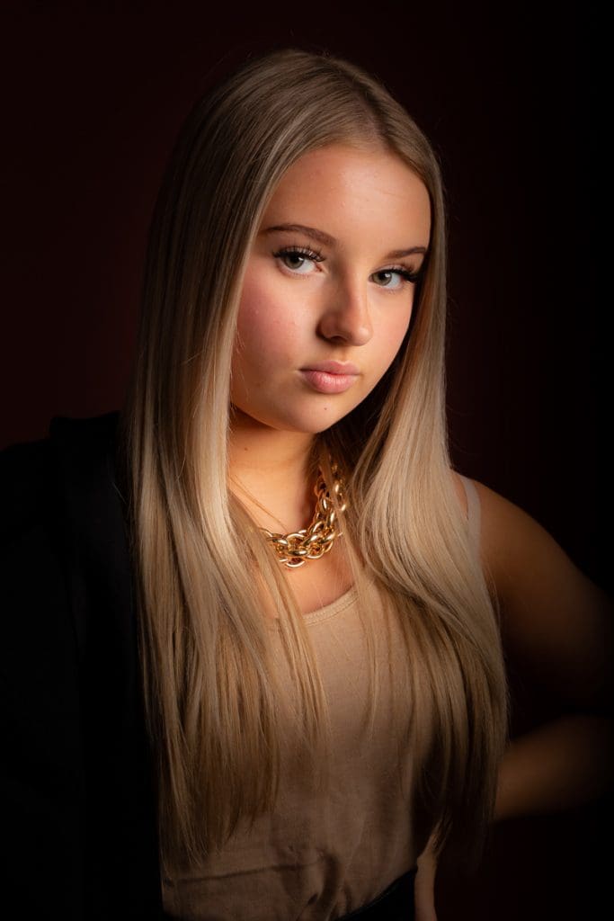 A young woman with long blonde hair poses against a dark background. She is wearing a beige top, a gold chain necklace, and a black jacket draped over her shoulders. Her expression is confident and calm.