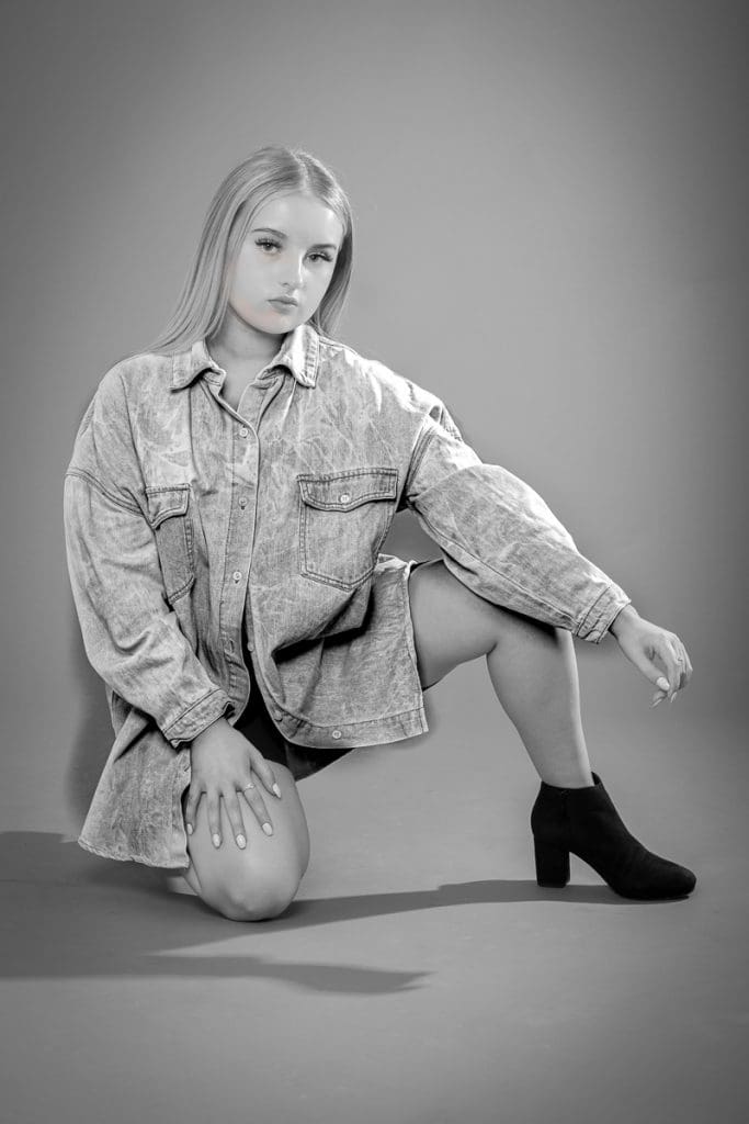 A person kneels on one knee against a plain backdrop, wearing an oversized denim shirt and dark ankle boots. The black and white image highlights the relaxed pose and confident expression.