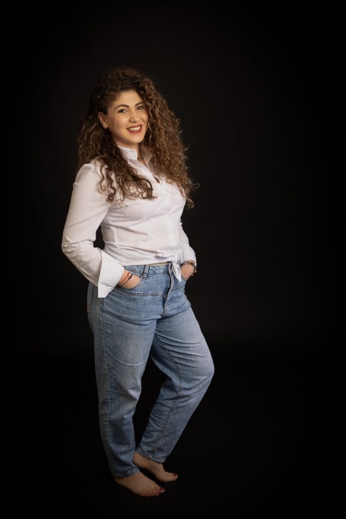 A person with curly hair, wearing a white blouse and blue jeans, stands barefoot against a dark background, smiling with hands in pockets.