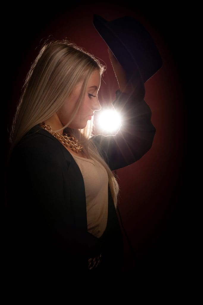 A woman with long blonde hair holds a hat above her head. She is in profile, wearing a black jacket and gold necklace. A bright light shines behind her, creating a halo effect. The background is dark.