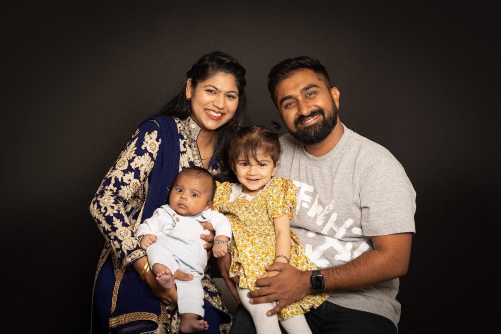 A smiling family of four poses together: a woman in blue, a man in a grey shirt, a young girl in a yellow dress, and a baby in white, all sitting closely against a dark background.