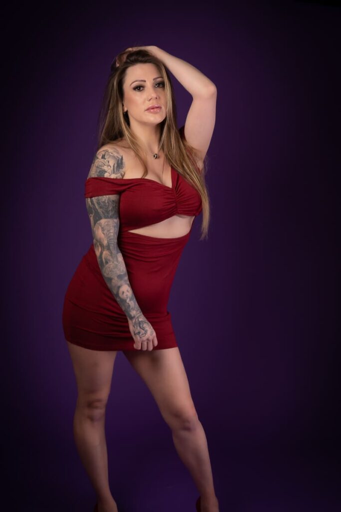 A woman in a red, off-the-shoulder dress with a cut-out stands against a dark purple background, one hand on her head. Captured by a Birmingham portrait photographer, she has long hair and a tattoo covering her left arm.