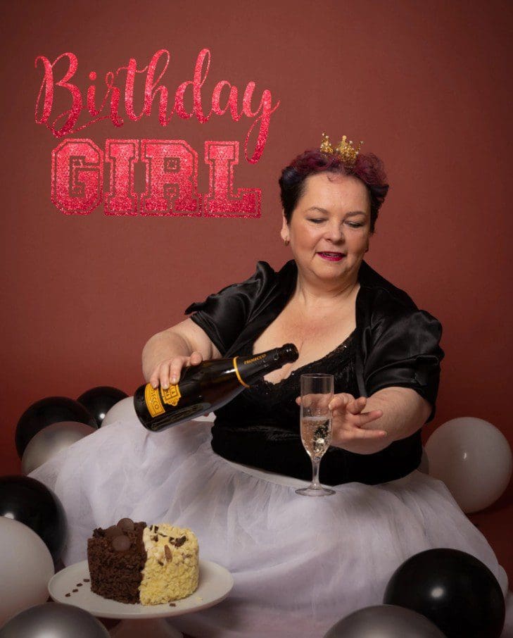 A woman in a crown and black top pours sparkling wine into a glass, with cake and black-and-white balloons around her, perfectly capturing the fun and glamour of a milestone celebration. Birthday Girl is written in pink glittery text above her—just one of the memorable moments styled by your Birmingham adult cake smash photographer.