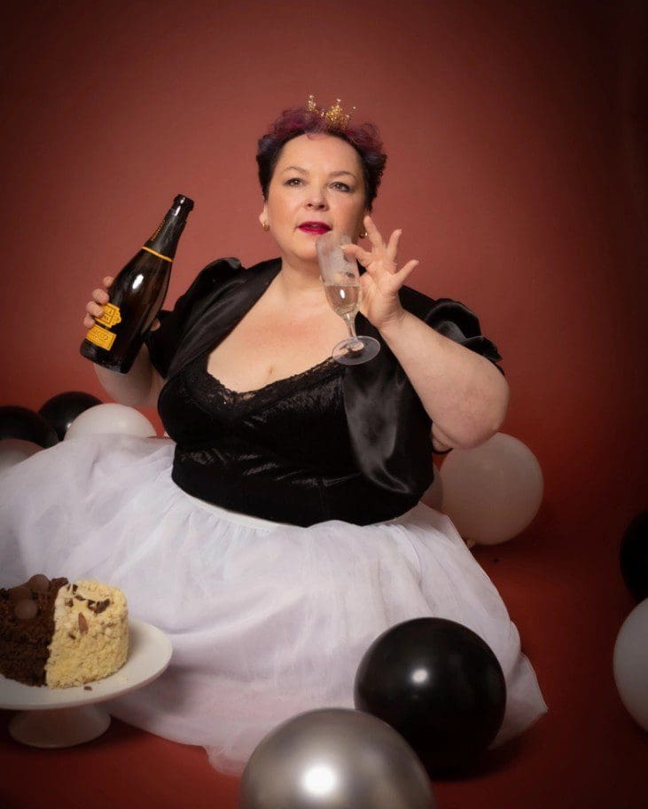 A woman in a black and white dress with a gold crown holds a champagne bottle and glass, sitting among balloons with a cake in front of her, against a reddish background—perfectly capturing the fun and elegance you’ll experience with a Birmingham adult cake smash photographer.