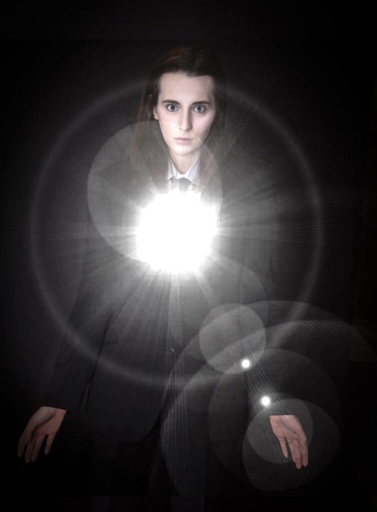 A person in a dark suit stands against a black background with a bright, glowing white light and lens flare effect shining from their chest, illuminating their face and hands—a striking image that captures the dramatic style often seen in Birmingham portrait photographer portfolios.