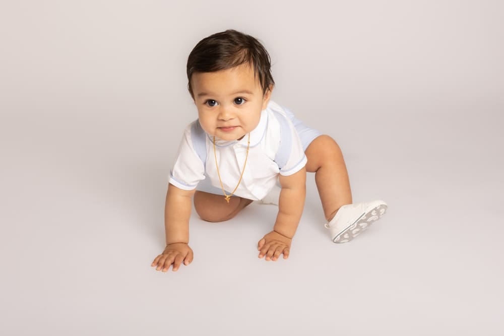 A baby wearing a white shirt, light blue shorts, and white shoes crawls on a light-coloured floor, looking up with a curious expression.
