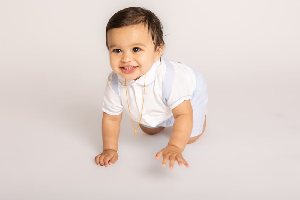 A smiling baby with dark hair wearing a white shirt, light blue shorts, and a gold necklace crawls on a plain white background.