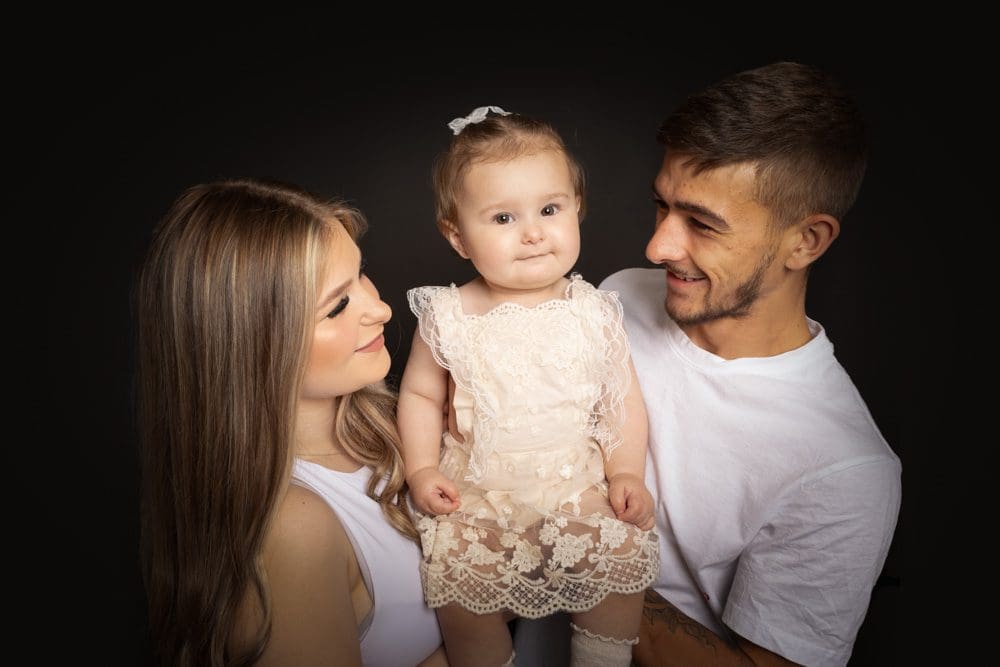 A smiling man and woman hold a baby girl dressed in a cream lace outfit. The adults look at the baby lovingly against a plain black background.