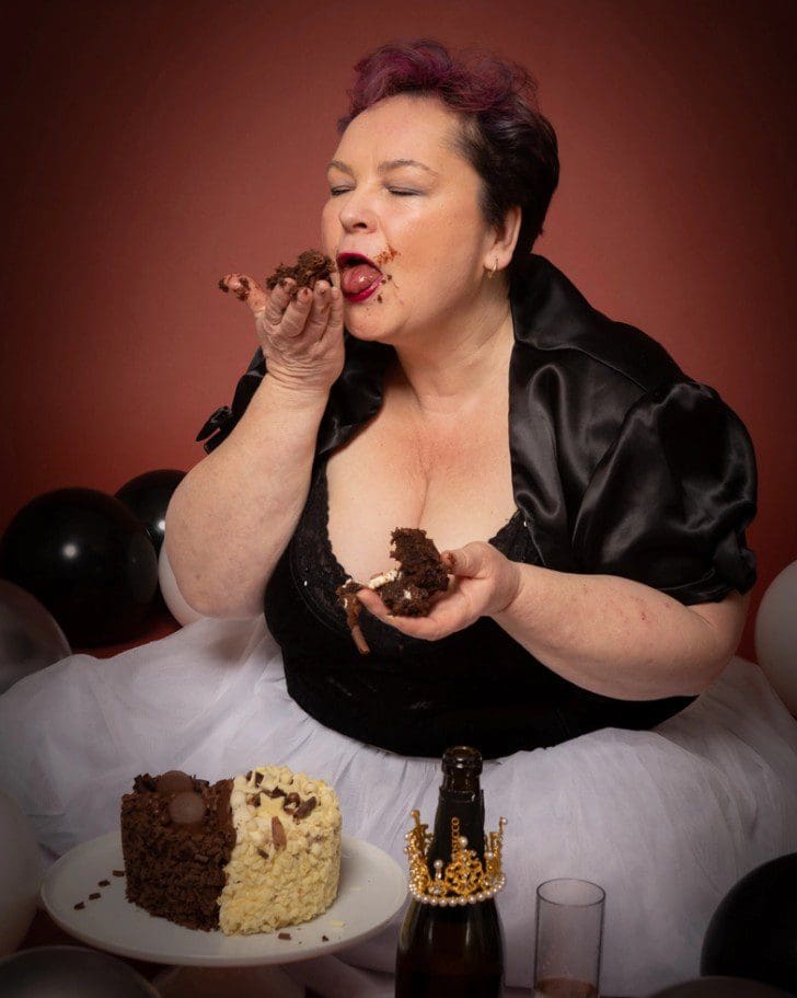 A woman in a black top and white skirt joyfully eats chocolate cake with her hands, surrounded by black and white balloons, a cake, and a bottle with a golden crown decoration—capturing the playful spirit of an adult celebration as seen through the lens of a Birmingham adult cake smash photographer.
