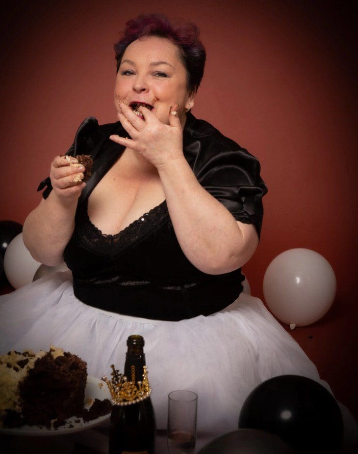 A person in a black top and white skirt enjoys eating cake with their hands, surrounded by black and white balloons, a cake, and a bottle on a table. The background is a warm reddish colour, creating the perfect fun-filled moment captured by a Birmingham adult cake smash photographer.