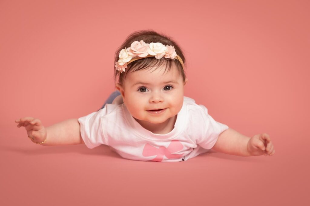 A smiling baby wearing a floral headband and white shirt lies on their stomach against a pink background, with arms outstretched.
