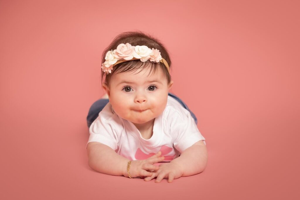 A baby wearing a floral headband and white shirt lies on their stomach against a pink background, looking forward with a slight smile.