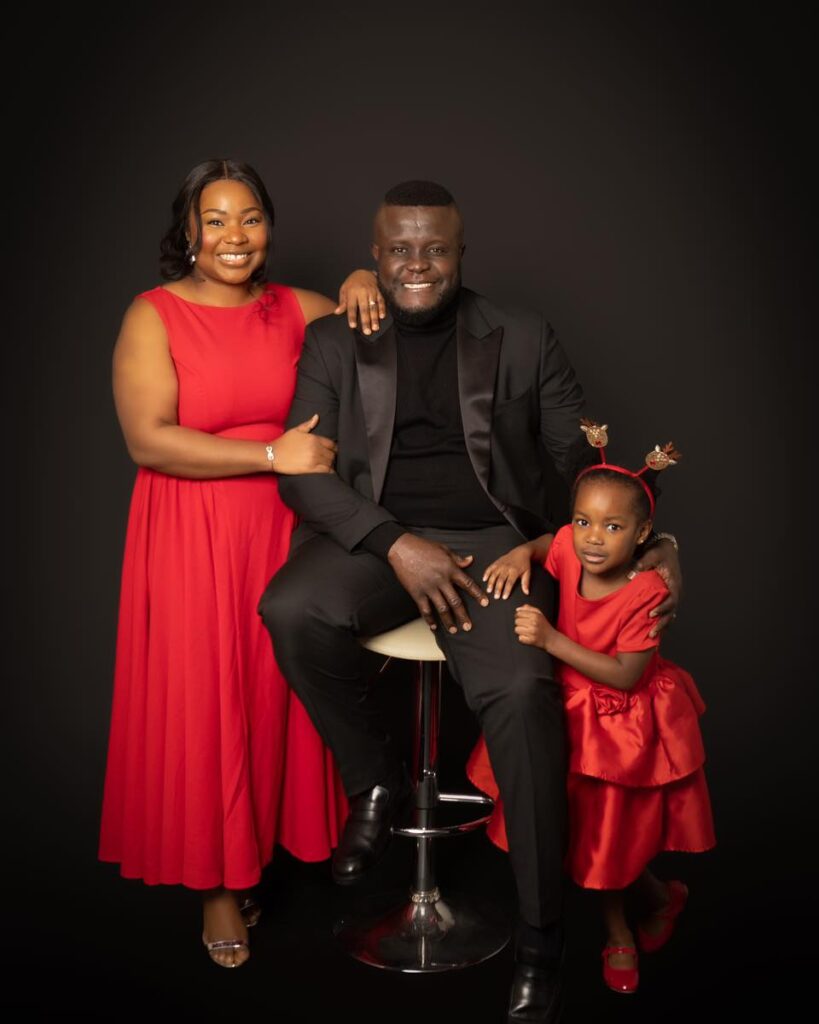 A smiling family poses against a black backdrop, perfectly capturing a moment that any Birmingham family photographer would cherish. A woman in a red dress stands on the left, a man in a black suit sits on a stool in the middle, and a young girl in a red dress sits on his knee, holding his hand.