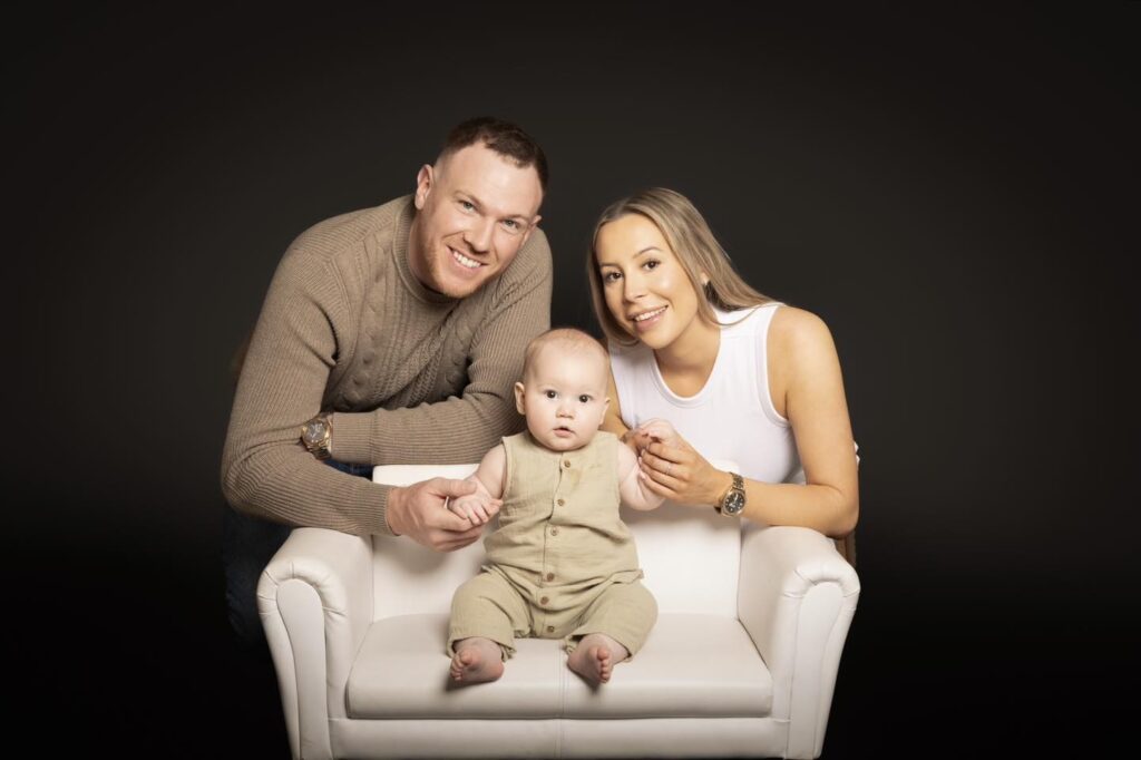 A smiling man and woman sit on either side of a baby who is seated on a small white sofa, all posing together against a dark background—a heartwarming moment beautifully captured by a Birmingham family photographer.