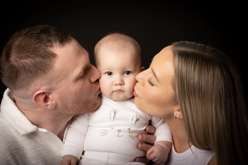 A baby in white is held between two adults who are kissing the baby’s cheeks from each side. The baby looks at the camera with a neutral expression, perfectly capturing a tender family moment often sought after by a Birmingham family photographer. The background is black.