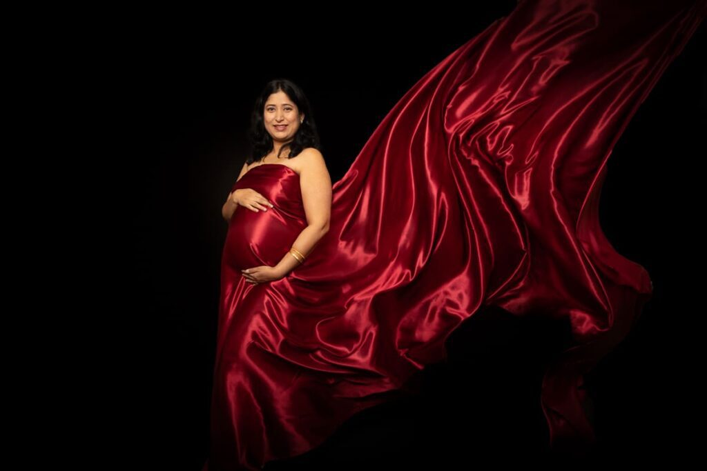 A pregnant woman in a flowing red satin dress poses against a black background, gently cradling her bump and smiling at the camera.