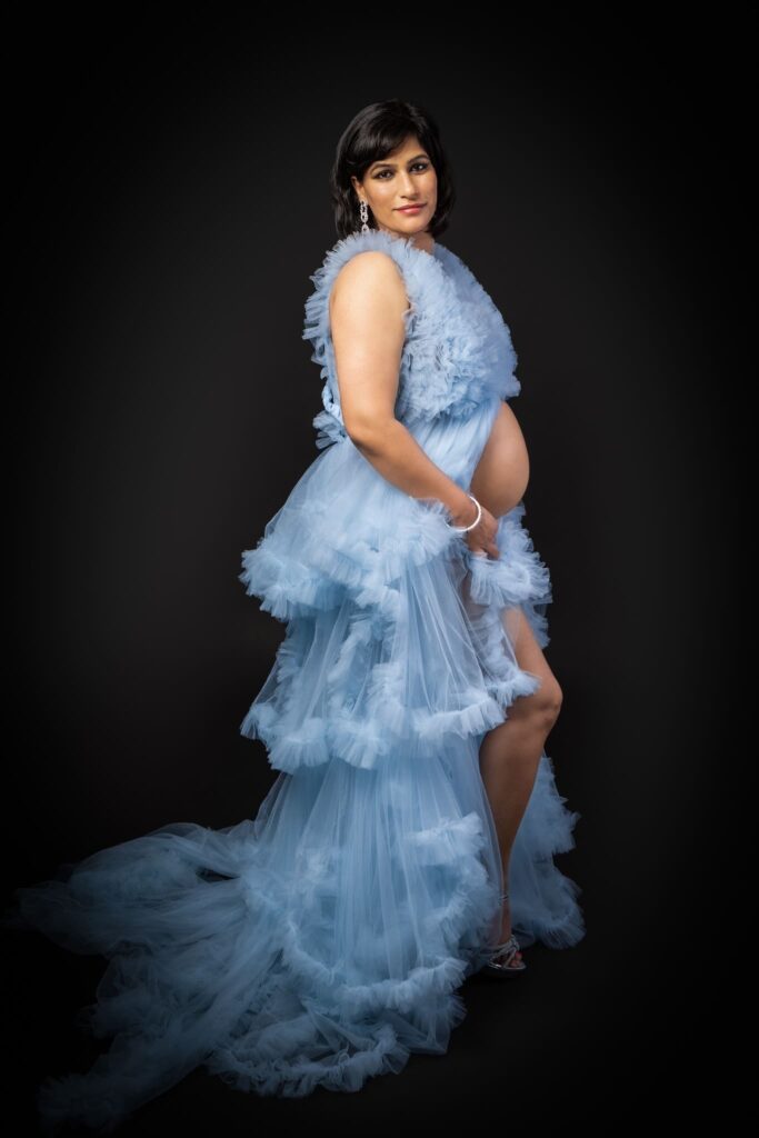 A pregnant woman in a light blue, ruffled gown poses against a black background, gently holding her exposed bump and looking at the camera.