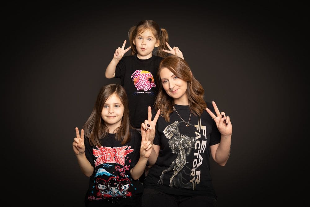 Three people, two children and a woman, pose against a dark background. All wear graphic T-shirts and hold up peace signs with their hands, smiling and looking at the camera—a joyful moment beautifully captured by a Birmingham family photographer.