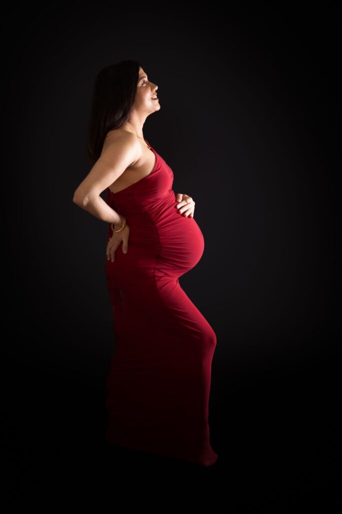 Pregnant woman in a long, fitted red dress stands in profile against a black background, smiling and holding her bump with one hand while the other rests on her lower back.