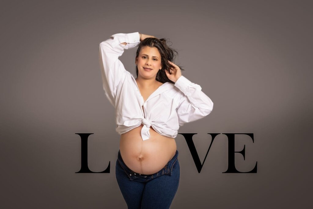 A pregnant woman in a white shirt and jeans poses confidently, standing in front of the word LOVE on a grey background.