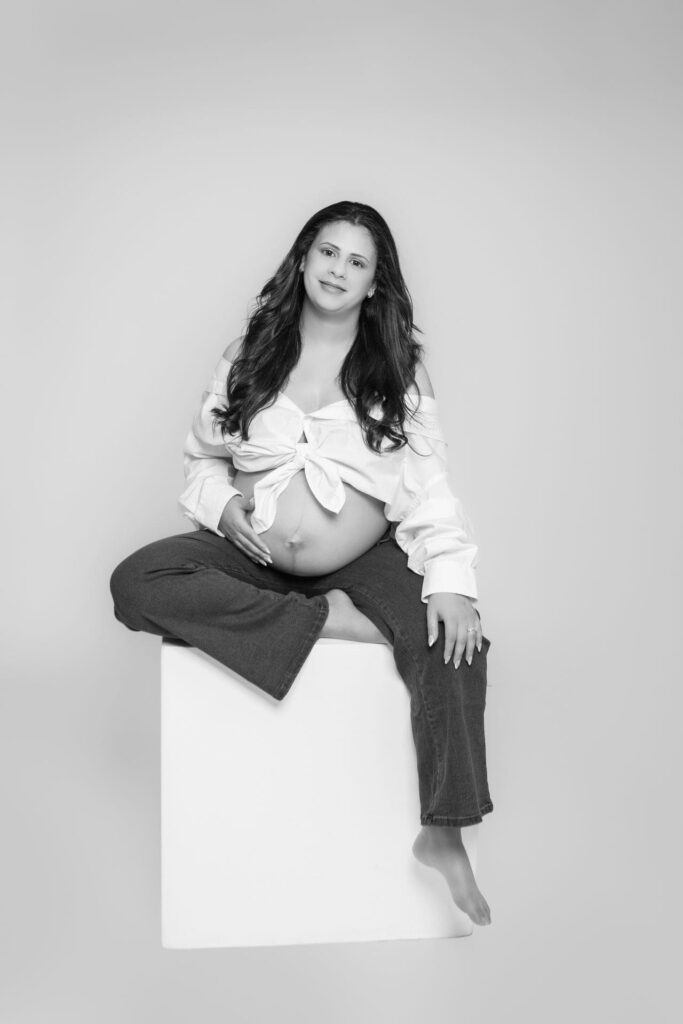 A pregnant woman sits barefoot on a white cube, wearing a tied white shirt and dark trousers, smiling softly and resting one hand on her bump. The photo is in black and white with a plain background.