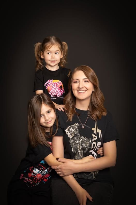 A woman sits smiling, with two young girls beside her. One girl stands behind with plaits, and the other leans in front. All are wearing black graphic T-shirts against a dark studio background, beautifully captured by a Birmingham family photographer.