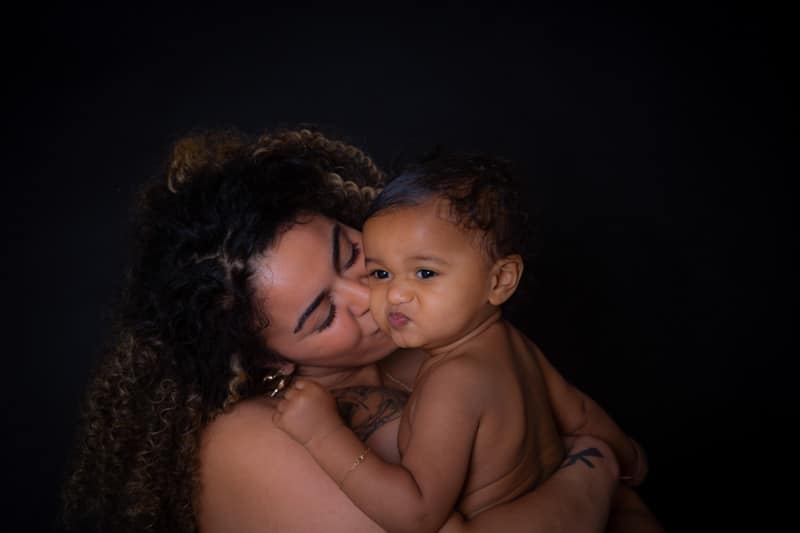 A woman with curly hair kisses a baby on the cheek. The baby, held in her arms, looks at the camera with a playful expression. Both are against a dark background.