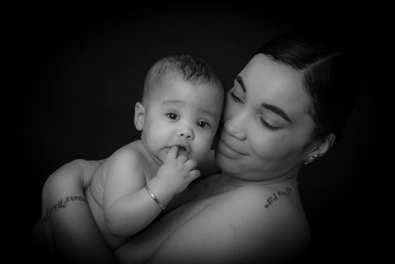 A woman smiles gently while holding a baby who looks at the camera with fingers in their mouth. Both are against a dark background.