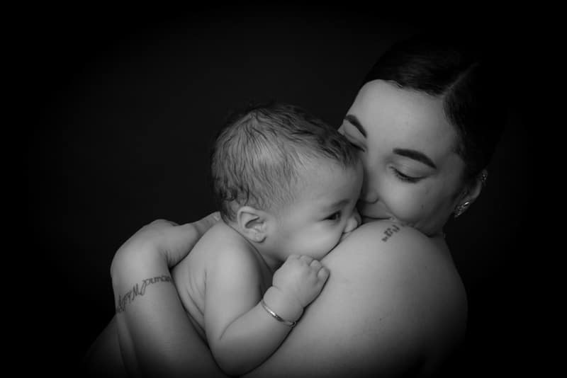 A black and white photo of a smiling woman hugging a baby close to her chest. The baby is nestled against her shoulder, and both have gentle, content expressions.