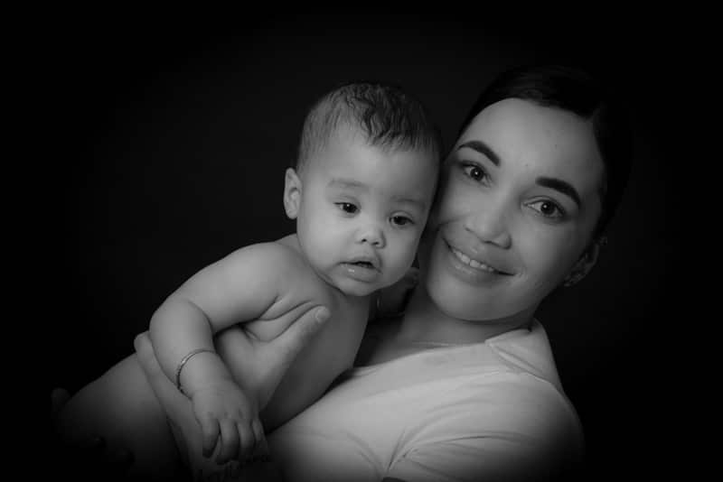 A woman smiles as she holds a baby in her arms. The photo is in black and white with a dark background, highlighting their faces.
