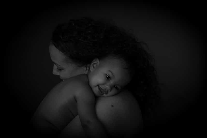 A black and white photo of a smiling baby resting on an adult’s shoulder. The adult’s face is partially hidden, while the baby looks at the camera, appearing comfortable and happy.