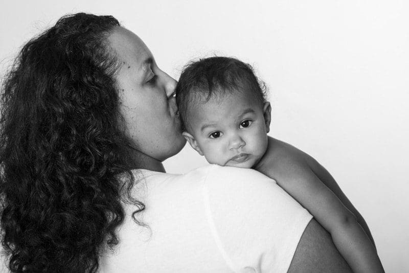 A woman with curly hair kisses a baby's head while holding the baby on her shoulder. The baby looks directly at the camera. The image is in black and white.