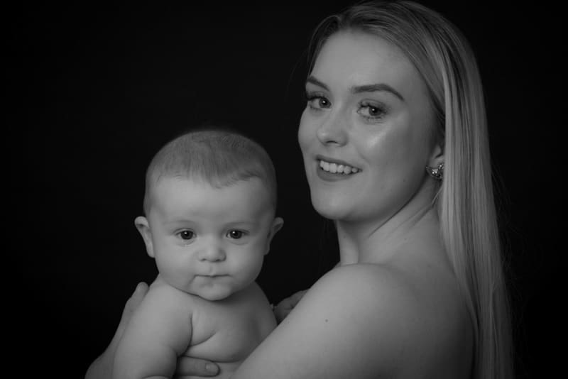 A woman with long hair smiles at the camera whilst holding a baby, both facing forwards against a dark background. The image is in black and white.