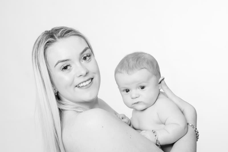 A woman with long blonde hair smiles whilst holding a baby in her arms. Both are looking at the camera against a plain white background.