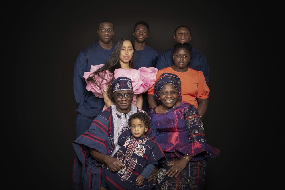 A family of eight poses against a dark background, dressed in colourful traditional and modern clothing. Two elders sit in front with a child, whilst five young adults stand behind them, each wearing different vibrant outfits.