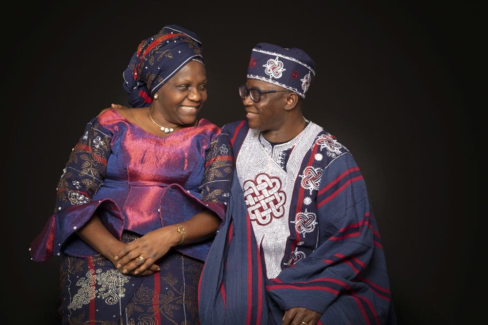 A smiling older couple in matching traditional African attire sit closely together against a dark background, looking at each other warmly.