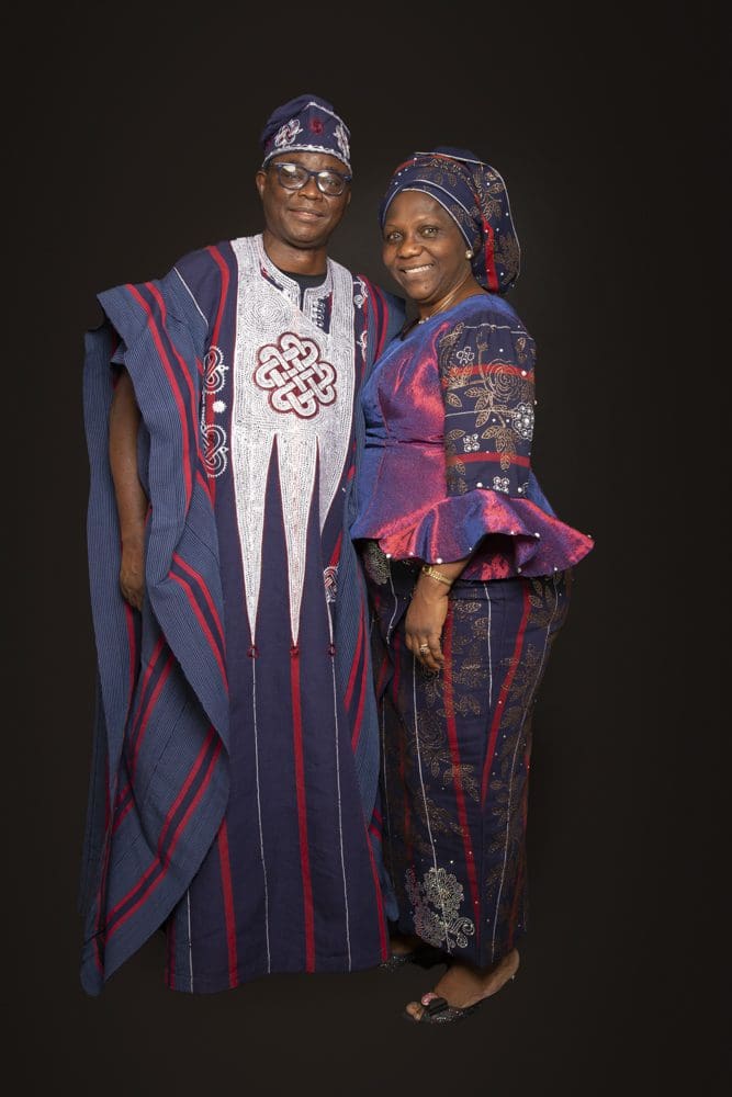 A man and woman stand smiling, wearing matching traditional Nigerian attire in blue, red, and white patterns, against a plain dark background.