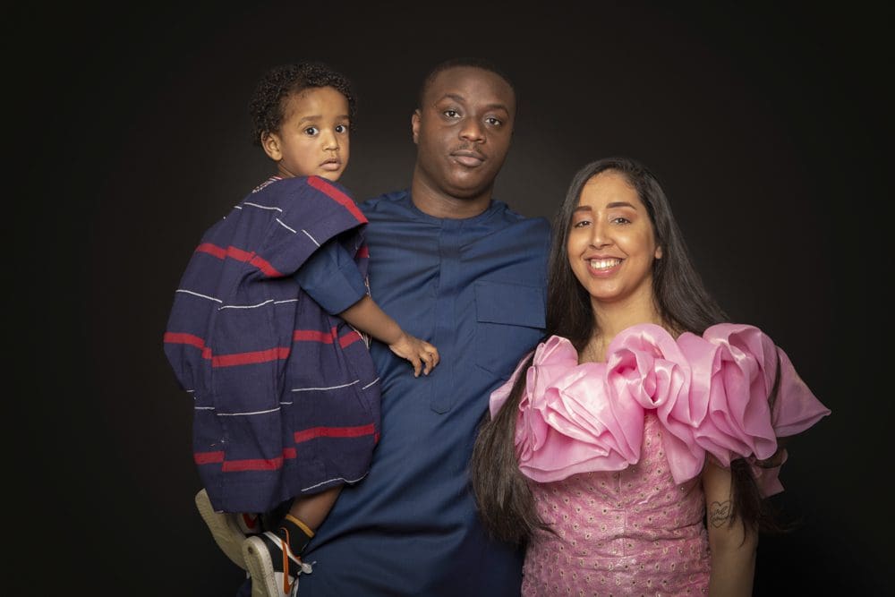 A man holding a young child stands next to a smiling woman wearing a dress with large pink frills, all posing against a plain dark background.
