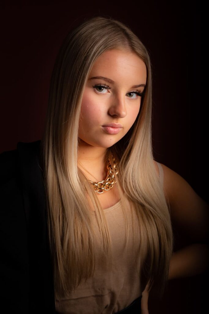 A young woman with long, straight blonde hair wears a beige top, a chunky gold chain necklace, and a black jacket draped over her shoulder, posing against a dark background in an image expertly captured by a Birmingham portrait photographer.