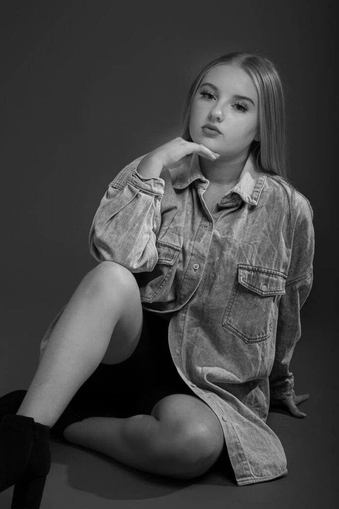 Black and white photo of a young woman sitting on the floor, wearing an oversized denim shirt and shorts, with one hand resting under her chin and looking thoughtfully at the camera—a striking image captured by a Birmingham portrait photographer.