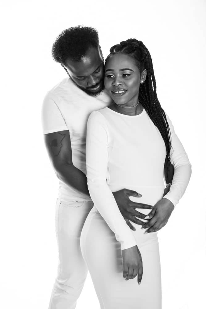 A man stands behind a woman, both dressed in white, smiling. The man gently places his hands on the woman's pregnant belly in a loving, joyful pose against a white background.