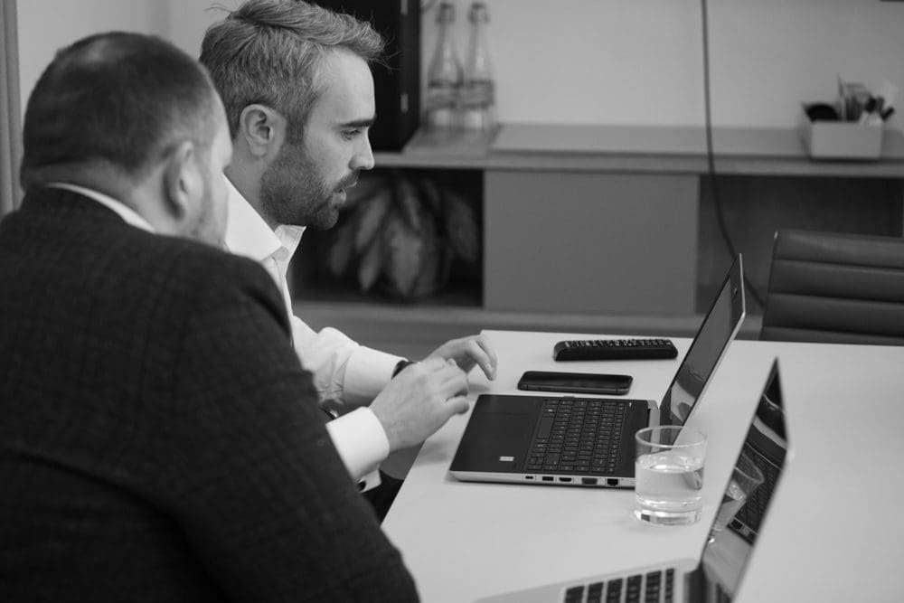 Two men in business attire sit at a table working on laptops, captured with the skilled eye of a Birmingham commercial photographer. On the table, there is a glass of water, a remote, and office supplies. Shelves and plants are visible in the background.