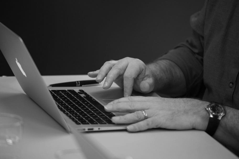 A person types on a laptop with one hand whilst the other hand rests nearby. A pen and a glass are visible on the table. Captured by a Birmingham commercial photographer, the image is in black and white.