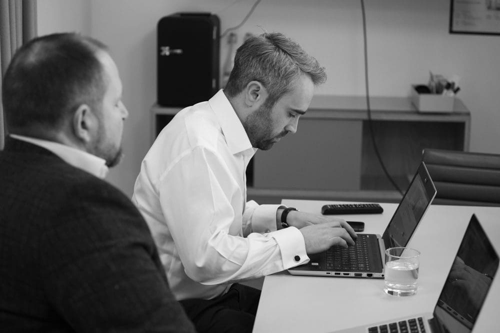 Two men in business attire sit at a table with laptops. One is typing, focused on his screen, whilst the other looks on. A glass of water and a remote control are also on the table. Captured by a Birmingham commercial photographer, this black and white photo exudes professionalism and concentration.
