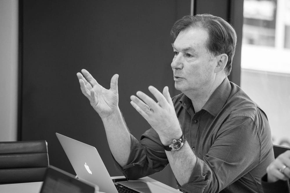 A man sits at a table with a laptop, gesturing with both hands as he speaks. Captured by a Birmingham commercial photographer, he appears engaged in conversation in a modern office setting.