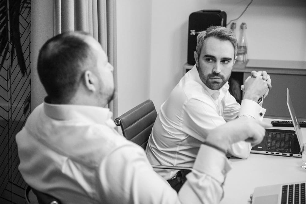 Two men in white shirts sit at a table with laptops, engaged in conversation. One faces the camera, looking intently at the other, who gestures with his hand. Captured by a Birmingham commercial photographer, the setting appears to be an office or meeting room.