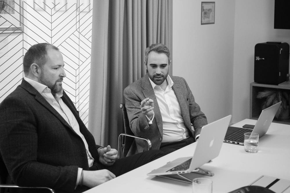 Two men in suits sit at a table with laptops, engaged in discussion. One gestures with his hand while the other listens, captured expertly by a Birmingham commercial photographer. Glasses of water and office décor are visible in the background. Black and white image.